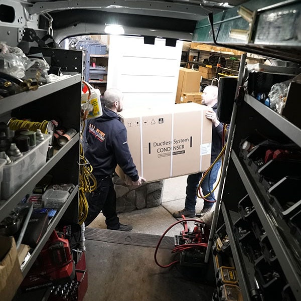 Two HVAC technicians carrying a box of new HVAC equipment out of their work van.